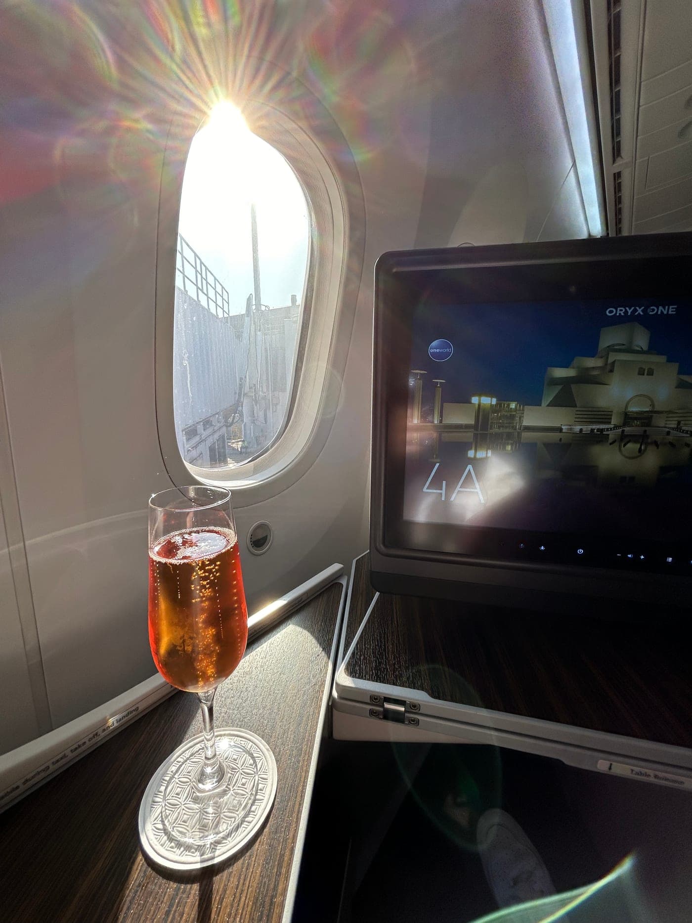 Fensterplatz 4A in der Business Class von Qatar Airways, ein Glas Champagner auf der Holzablage, auf dem Bildschirm die Begrüßung mit dem Ziel Doha