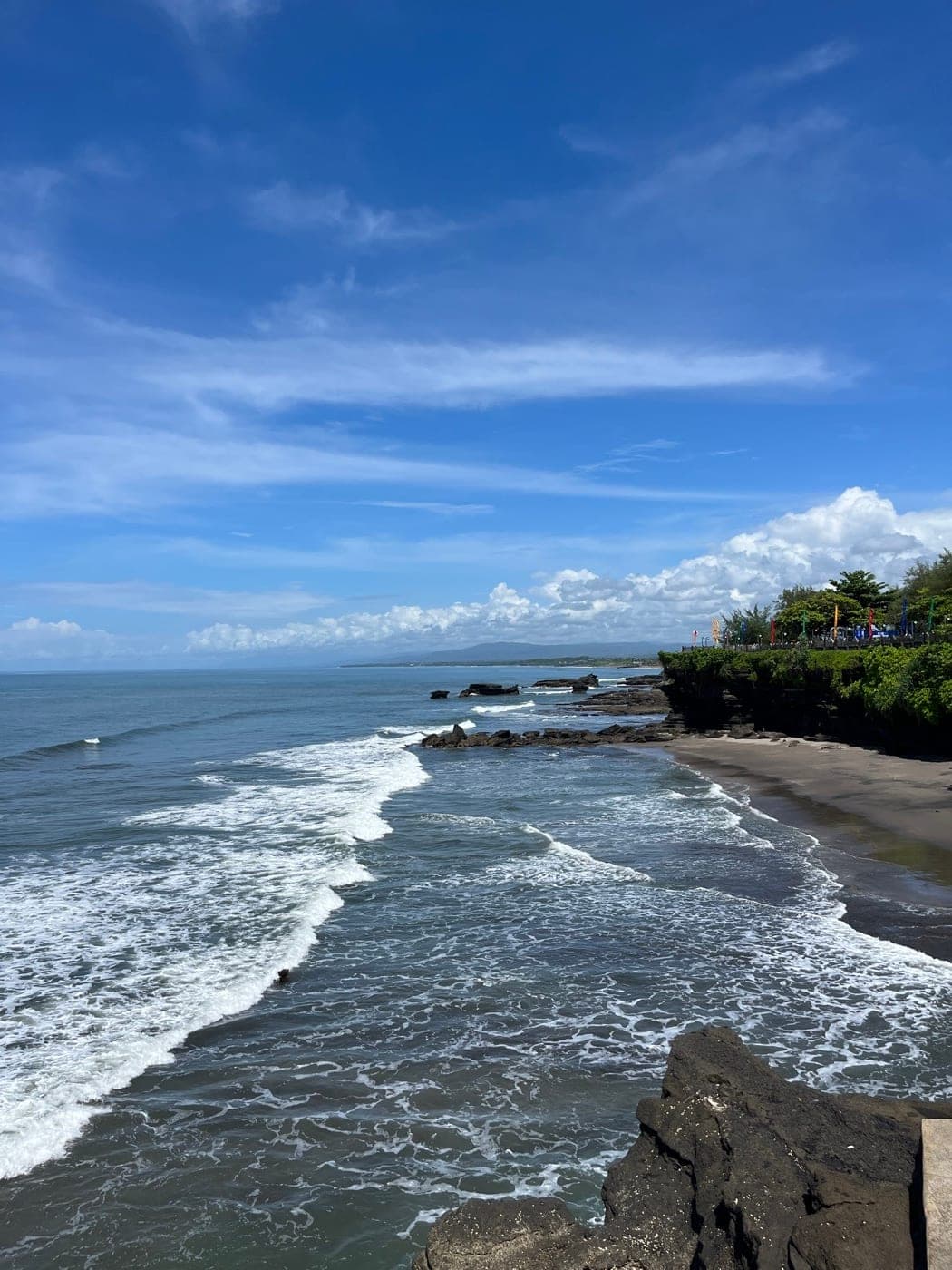 Kueste von Bali mit Brandung, dunklem Sandstrand und begruenter Klippe
