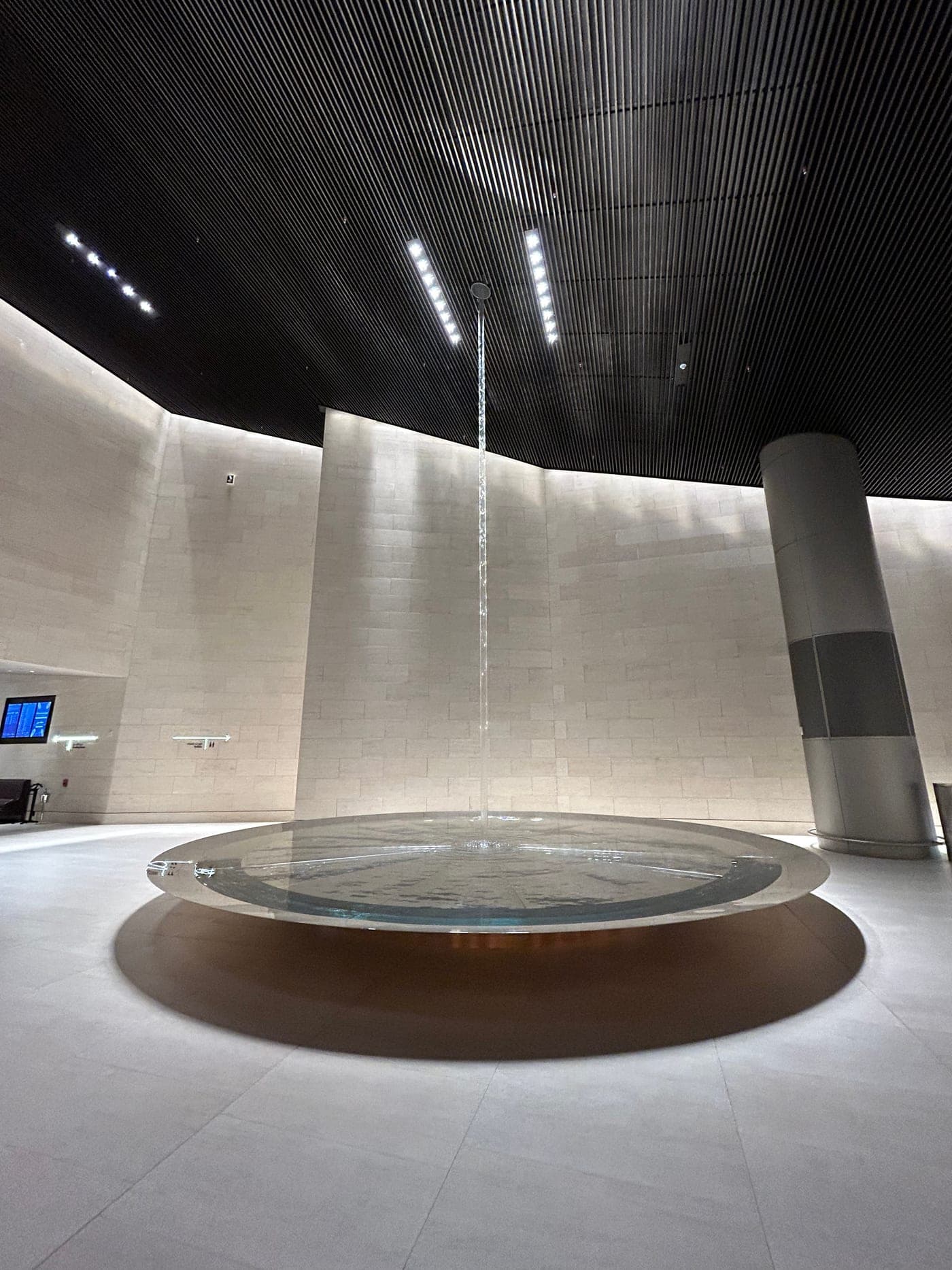 Runde Wasserinstallation in der Al Safwa Lounge: ein Wasserstrahl fällt von der dunklen Decke in ein flaches, beleuchtetes Becken